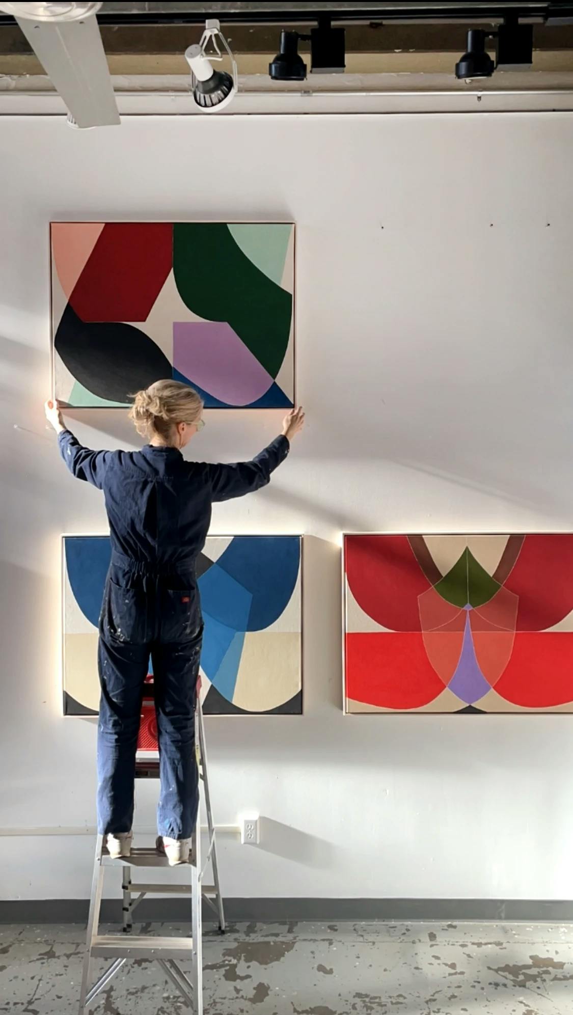 Artist Christina Flowers on a ladder in her studio holding up a large, abstract painting.