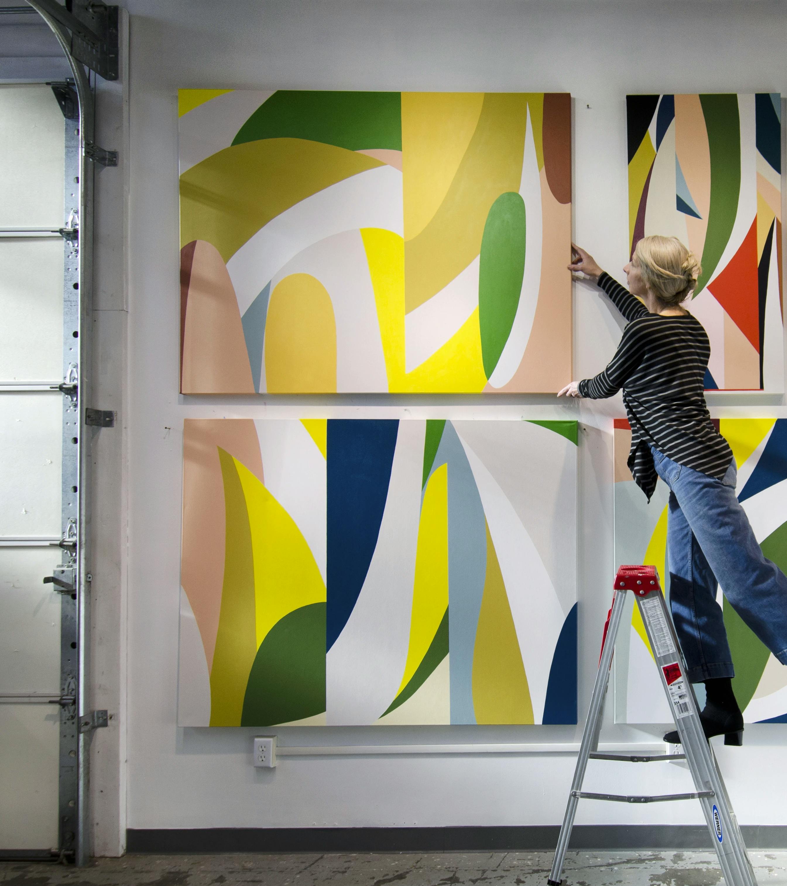 Artist Christina Flowers on a ladder installing a large, geometric abstract painting on a white wall in her studio.
