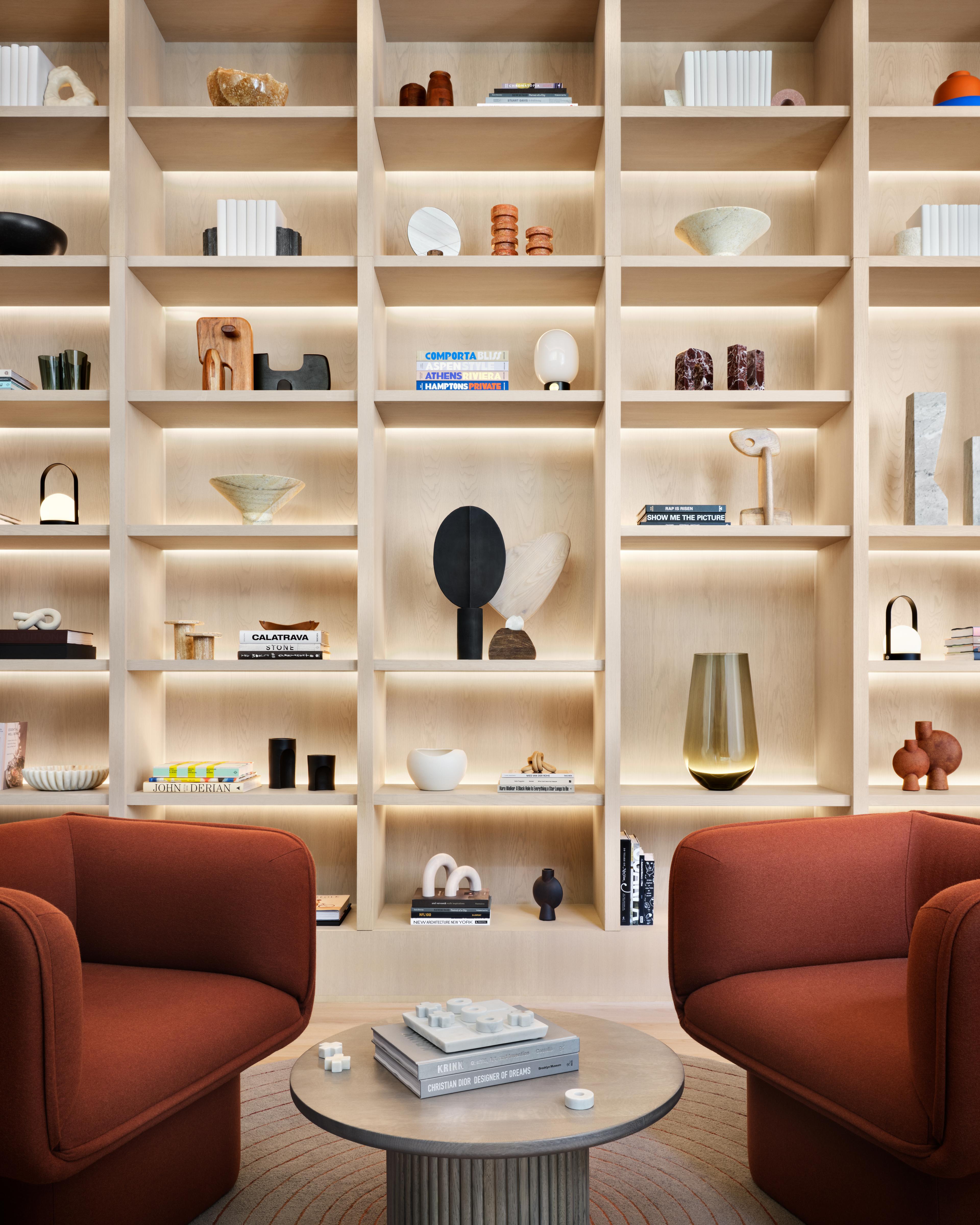 Wooden built-in bookshelves with various books, sculptures, and items within a lobby area with orange armchairs within Anagram NoMad.