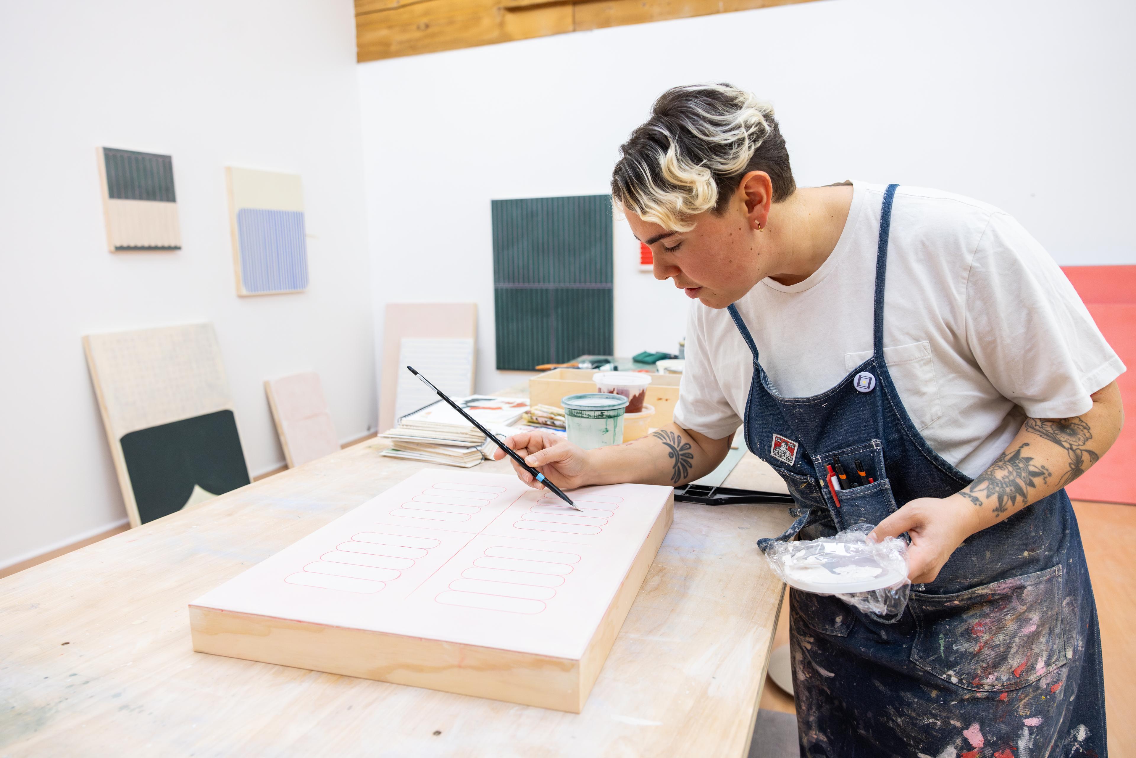 Artist Arielle Zamora painting a pink work on panel in their studio.