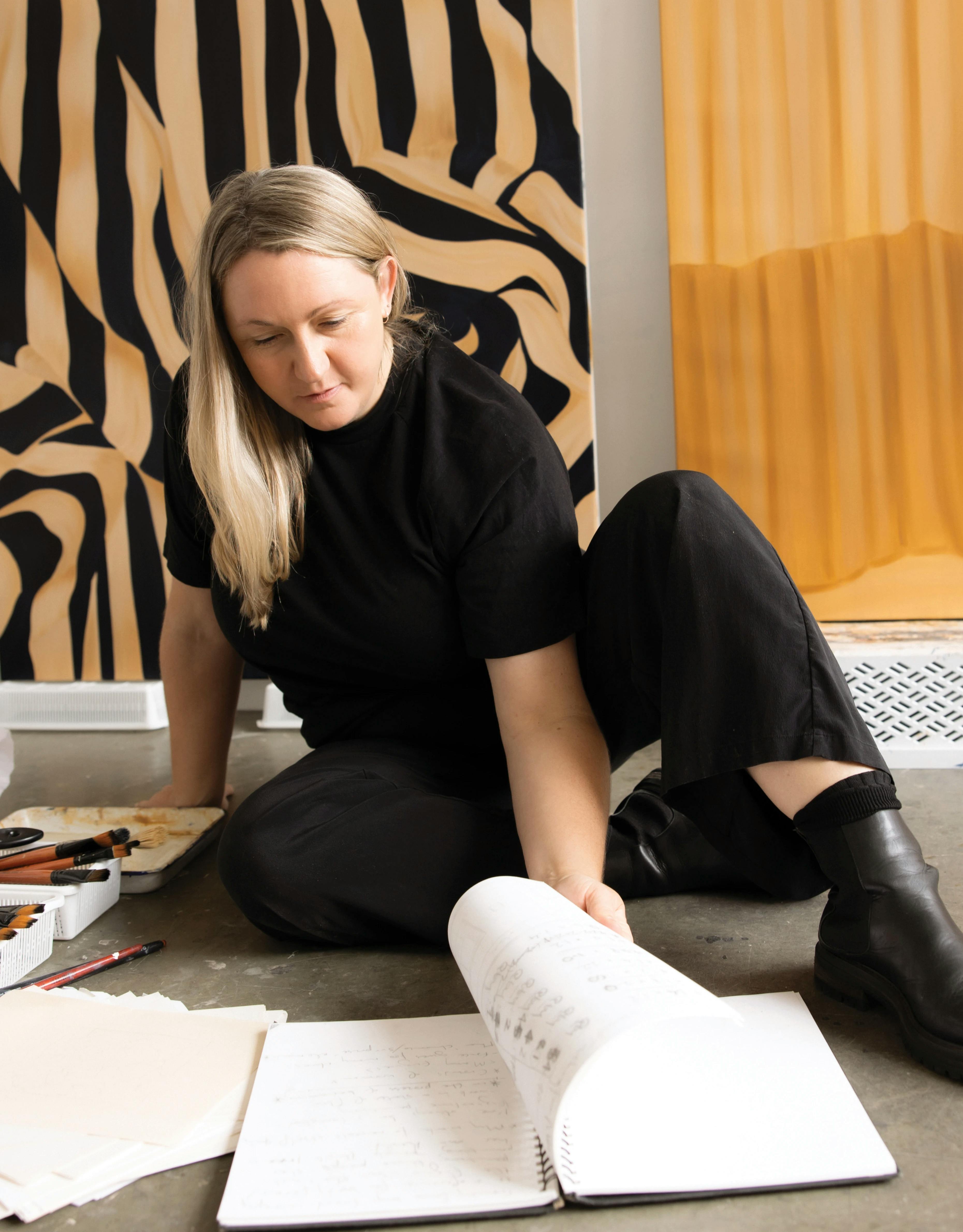 Artist Caroline Walls sitting on the floor of her studio flipping through a notebook.