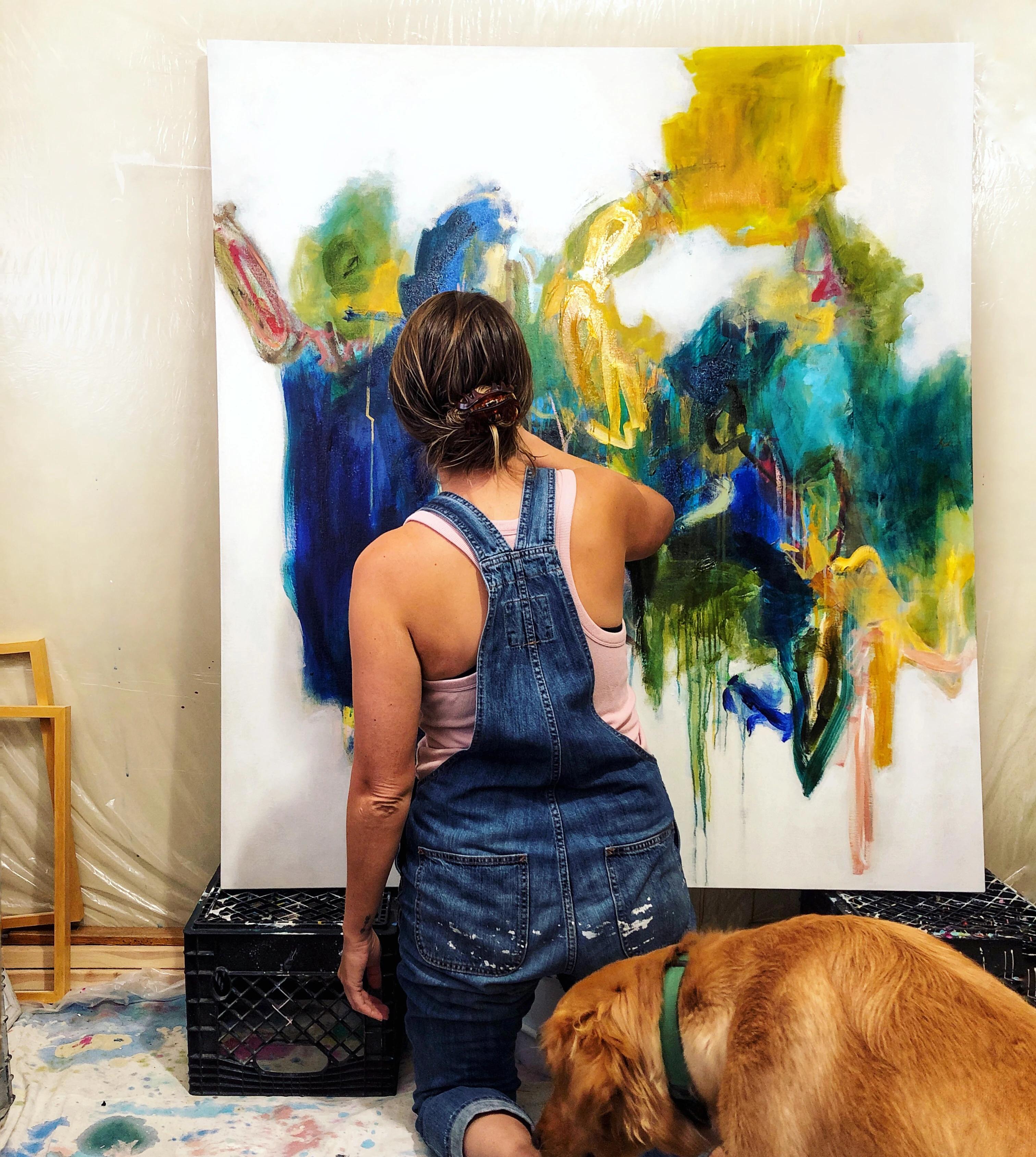 Artist Elisa Gomez kneeling next to her golden retriever dog, working on a large, blue and yellow abstract painting in her studio. 