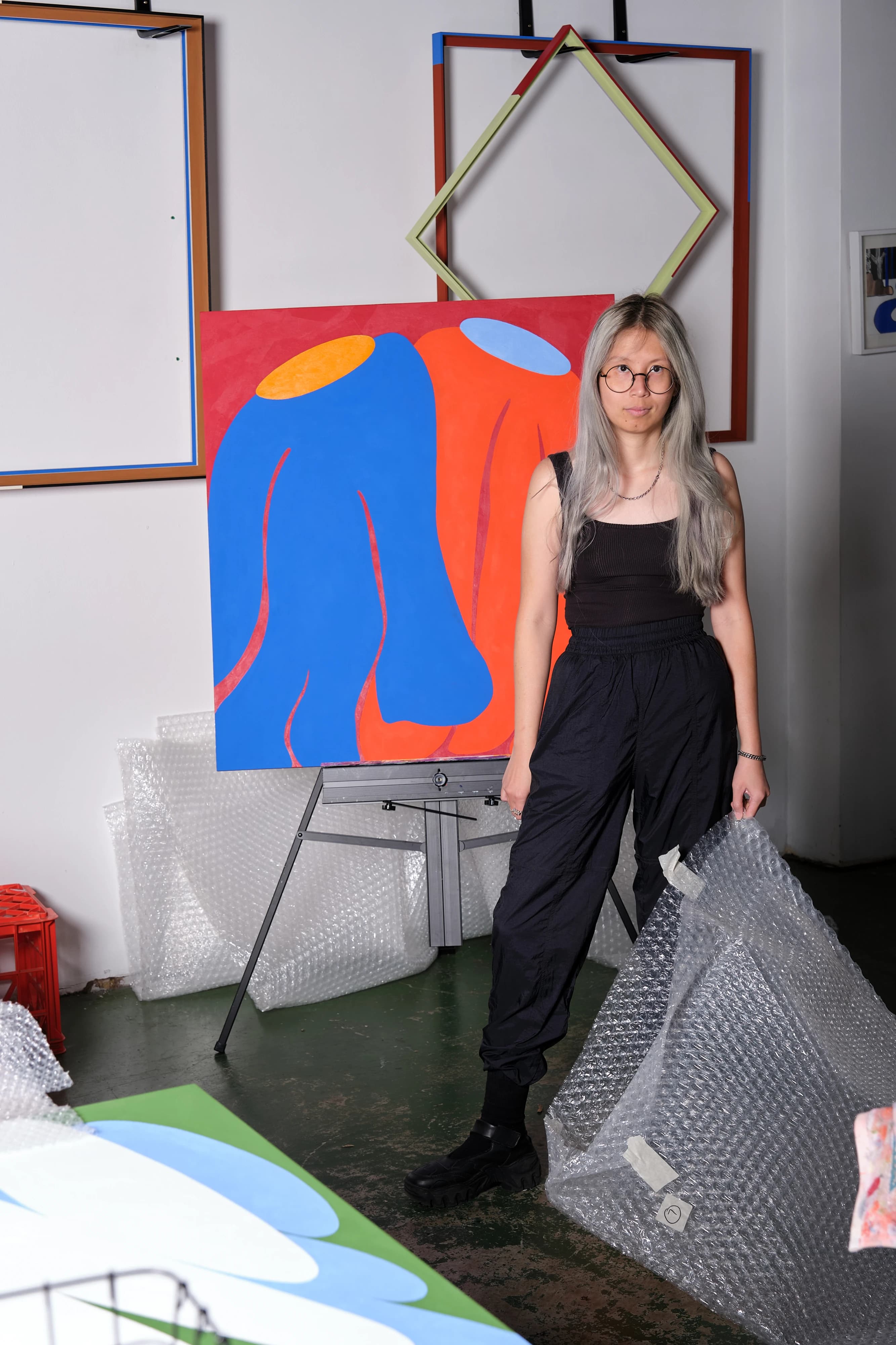 Artist Evi O. standing in front of a painting on an easel with amorphous red and blue shapes.