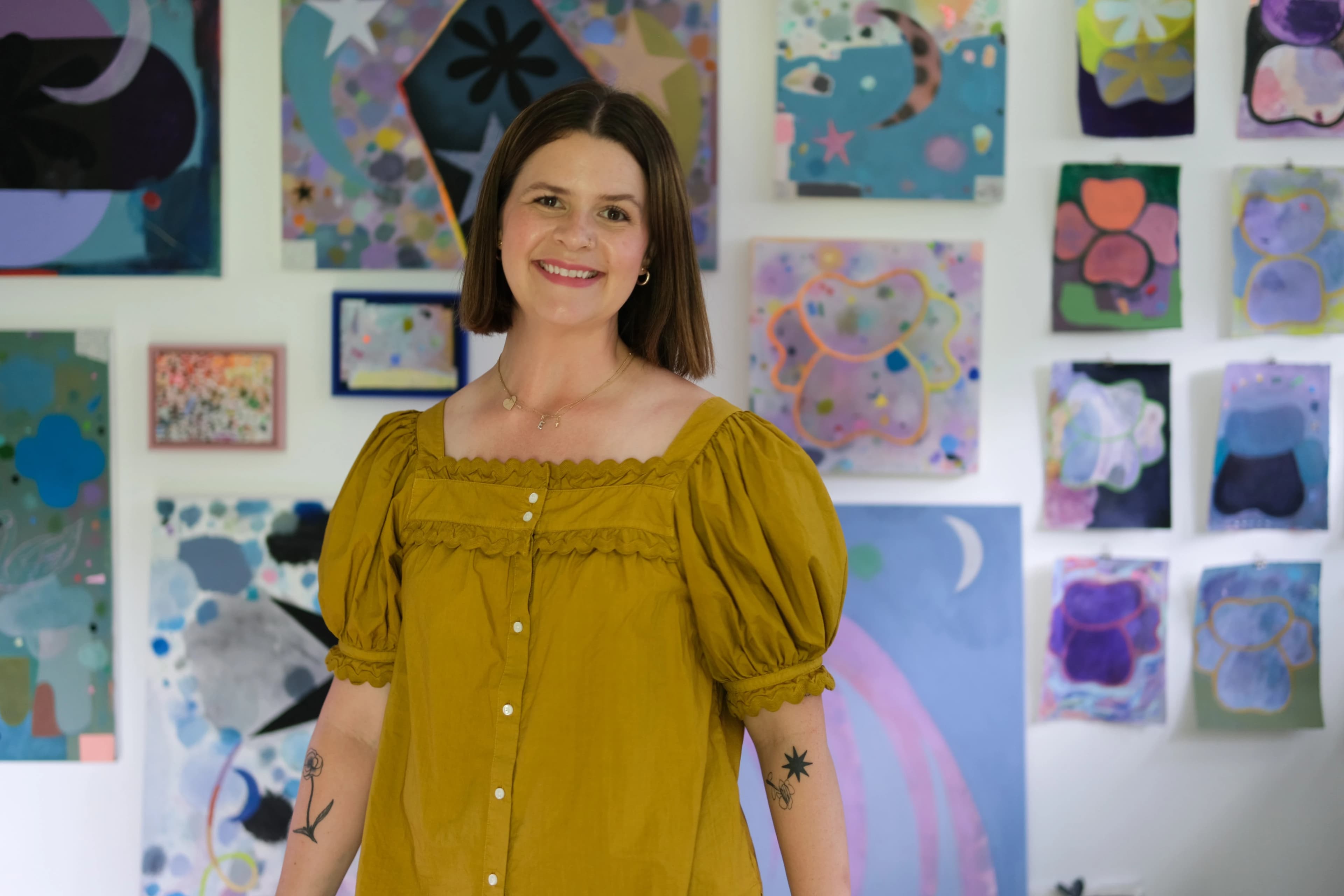 Artist Ashely Peifer wearing a mustard yellow shirt in her studio, surrounded by her painting installed on a white wall behind her.