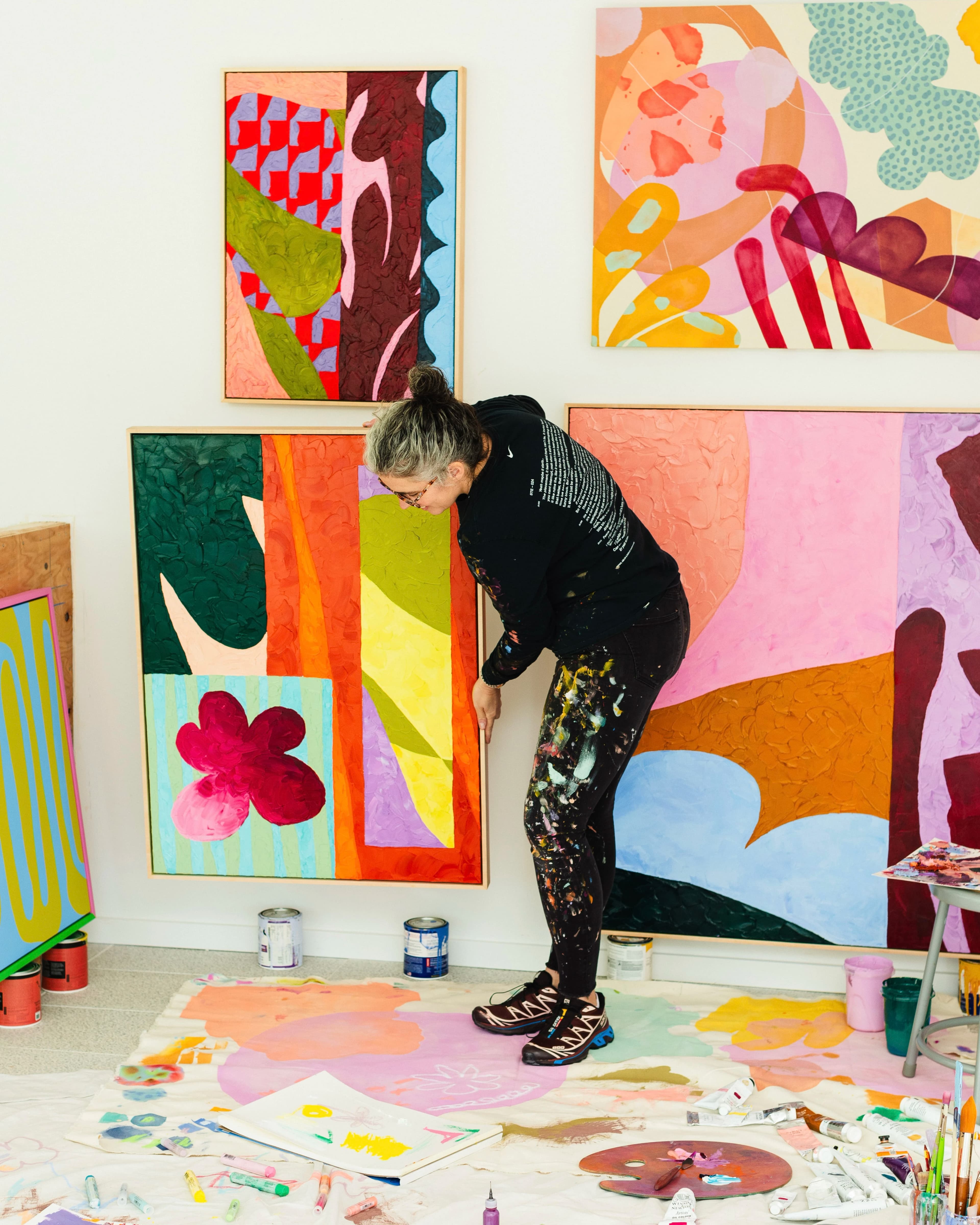 Artist Alex Proba holding a brightly colored, abstract canvas in her studio surrounded by her other vibrant works.