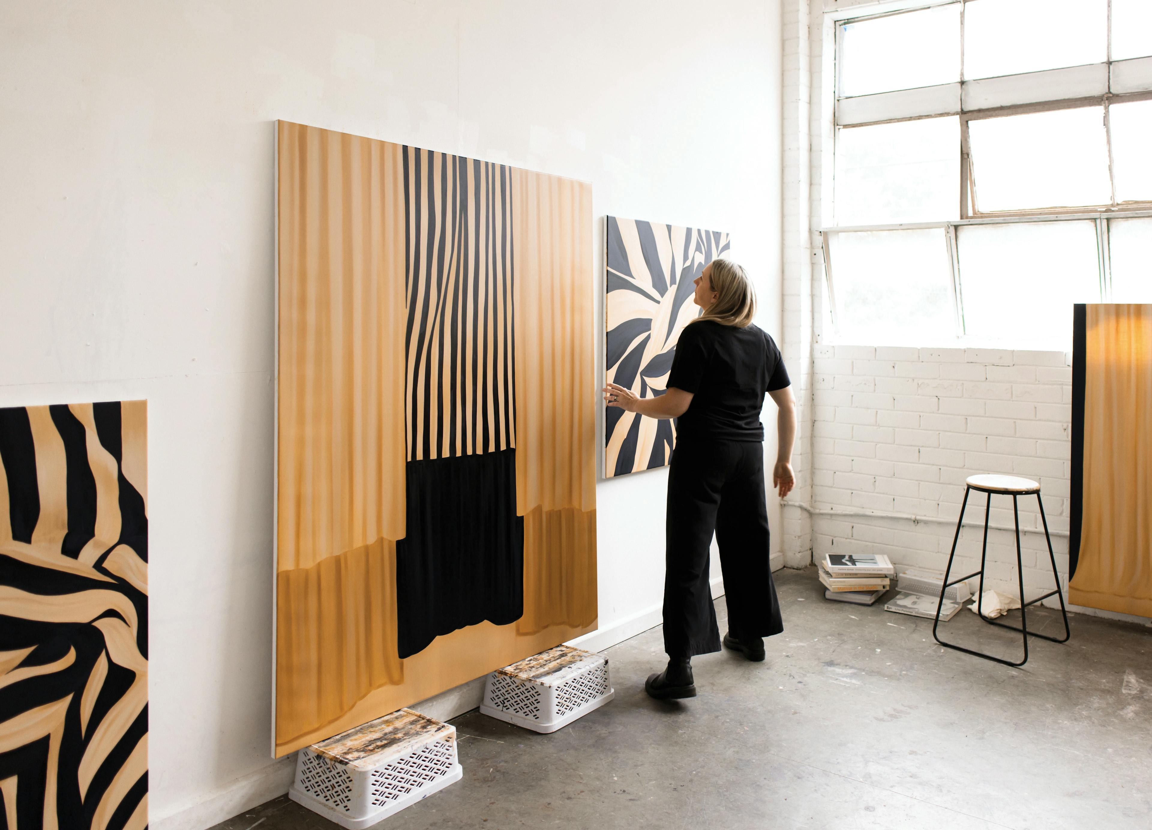 Artist Caroline Walls in her studio with large, beige and black striped paintings on canvas.