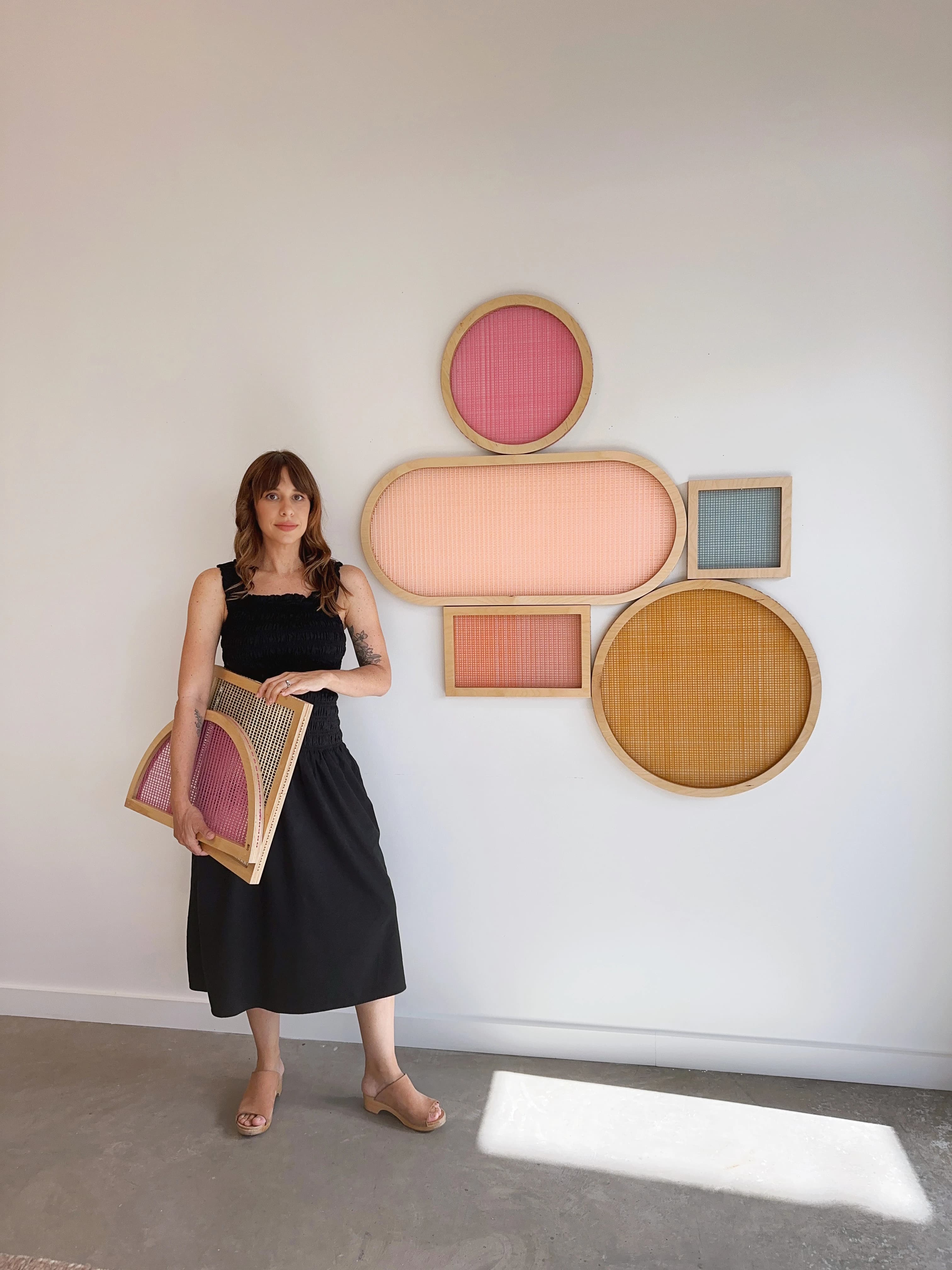 Artist Hayley Sheldon holding two colorful screens, standing next to artwork installed on the wall.