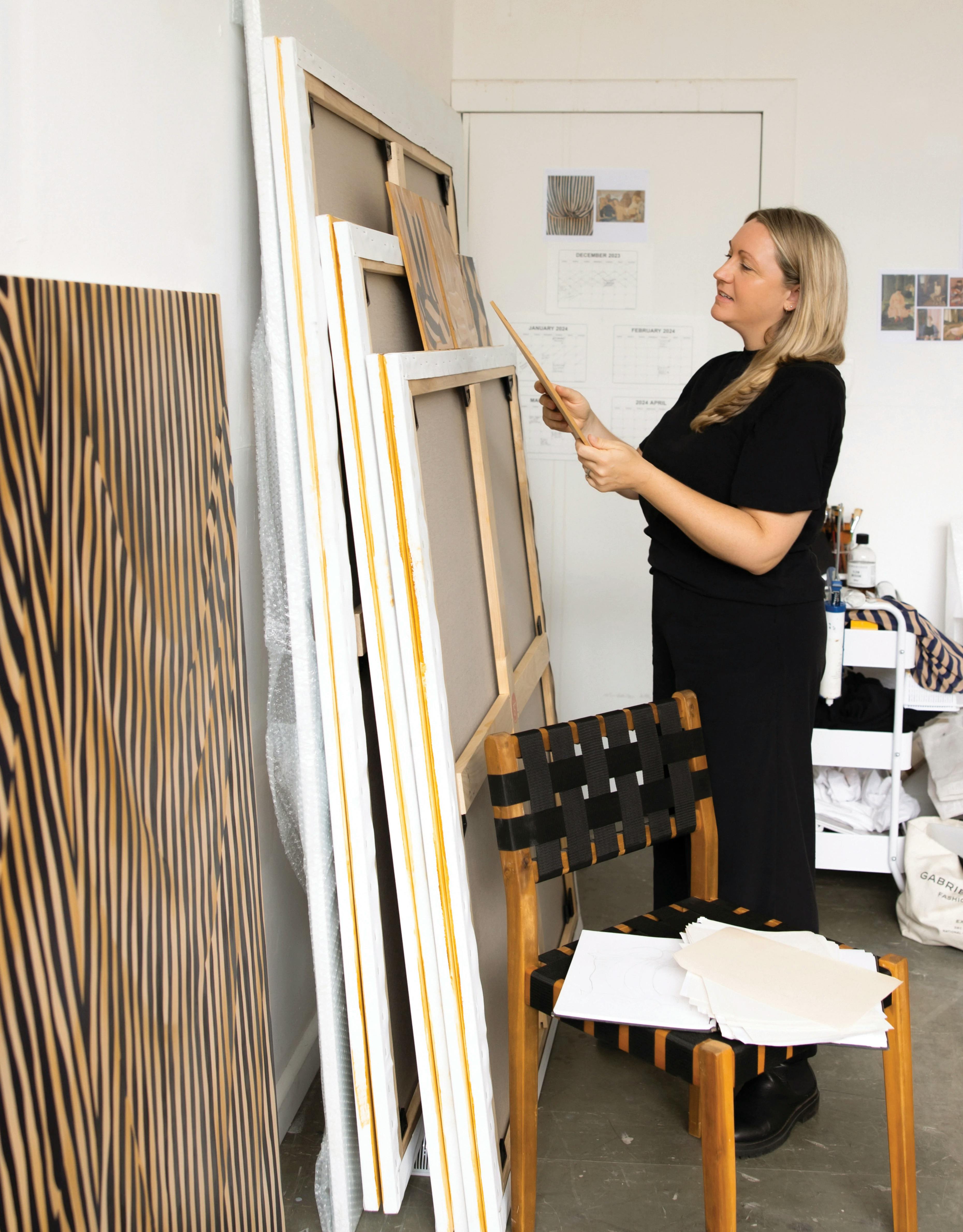 Artist Caroline Walls standing in her studio next to a stack of paintings on canvas.