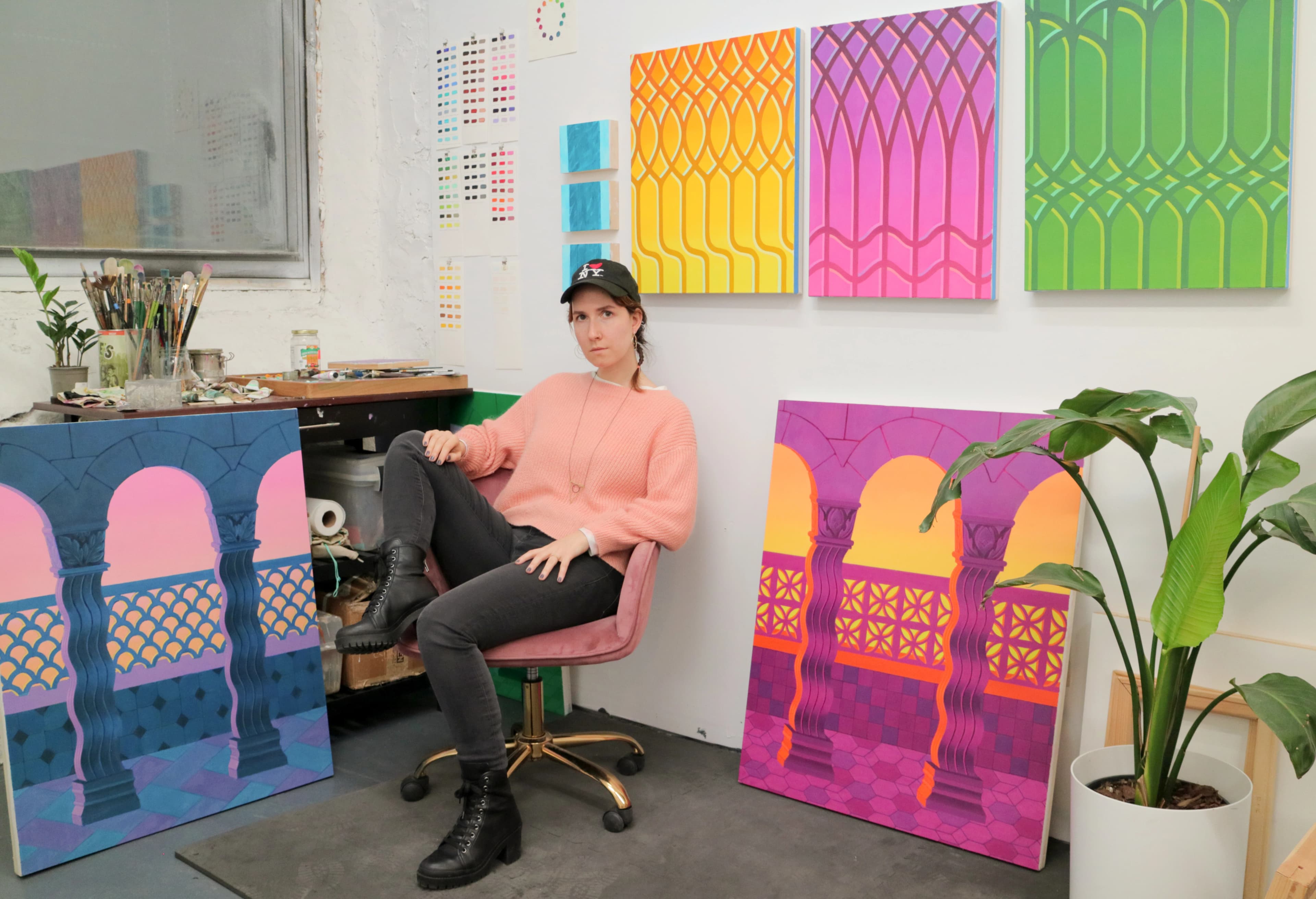 Artist Lucía Rodríguez Pérez sitting in a chair in her studio surrounded by her colorful, architectural and botanical paintings.