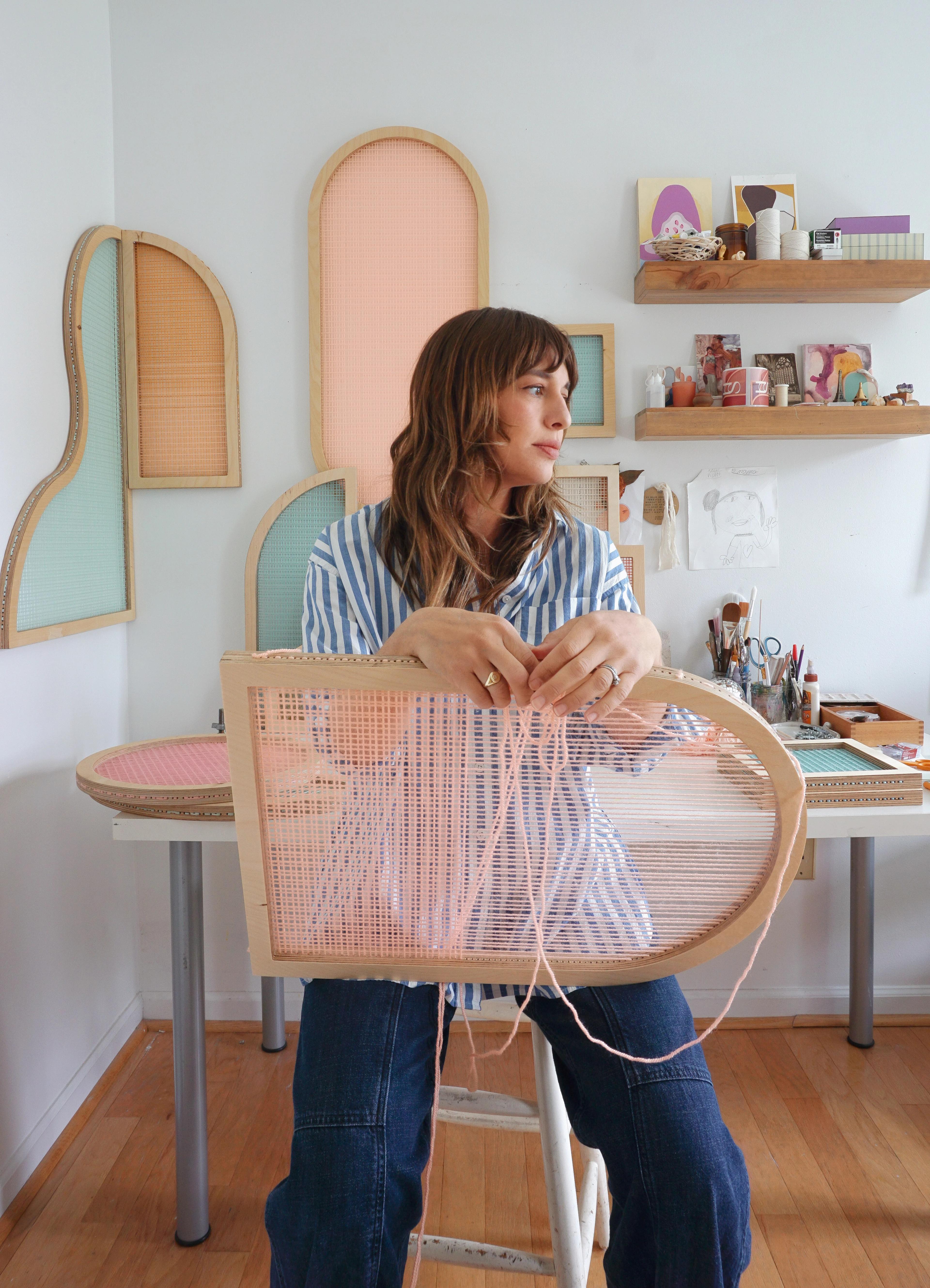 Artist Hayley Sheldon sitting in her studio holding a light pink arched shape screen.