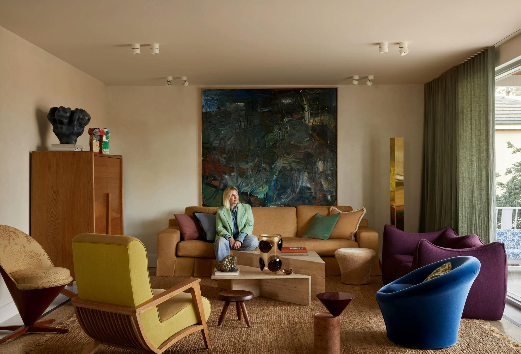 Designer Tali Roth sitting on a brown couch below a large abstract painting within a living room with colorful chairs.