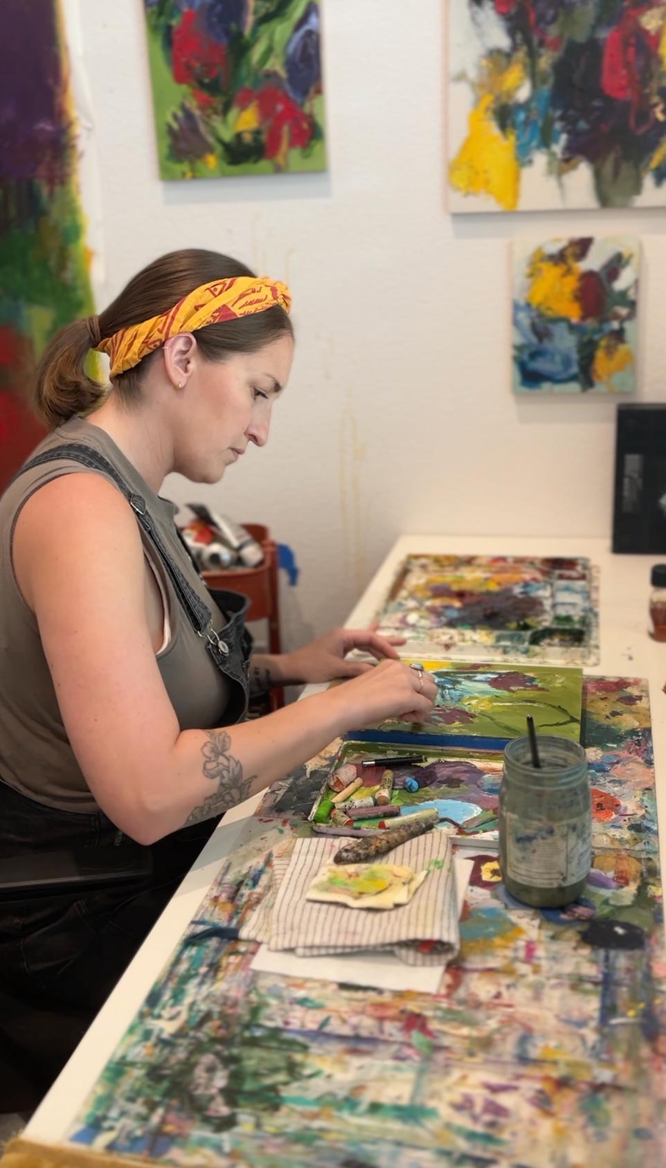 Artist Elisa Gomez working on a small, colorful painting at a table in her studio.