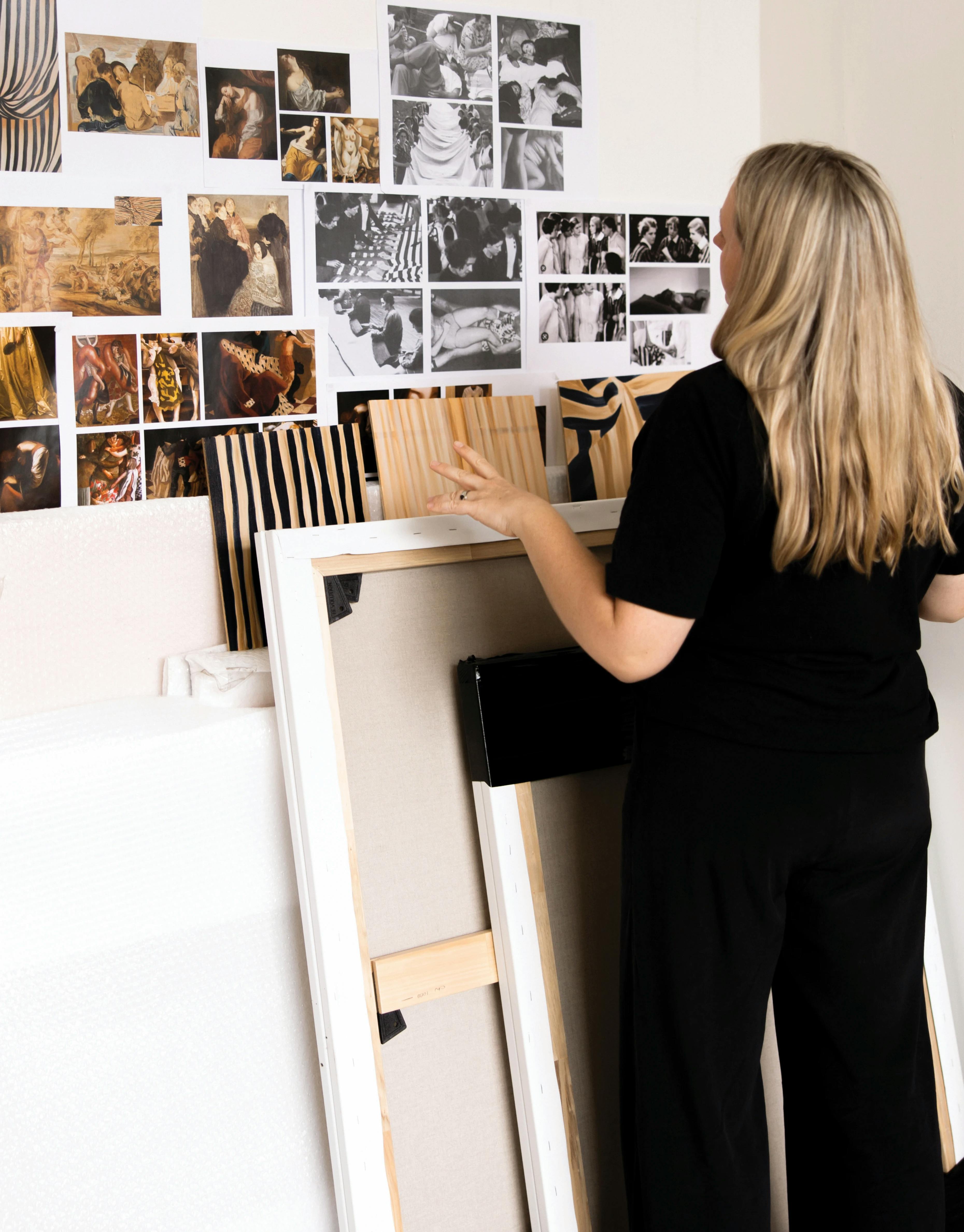 Artist Caroline Walls standing in her studio looking at photographs taped to the wall.