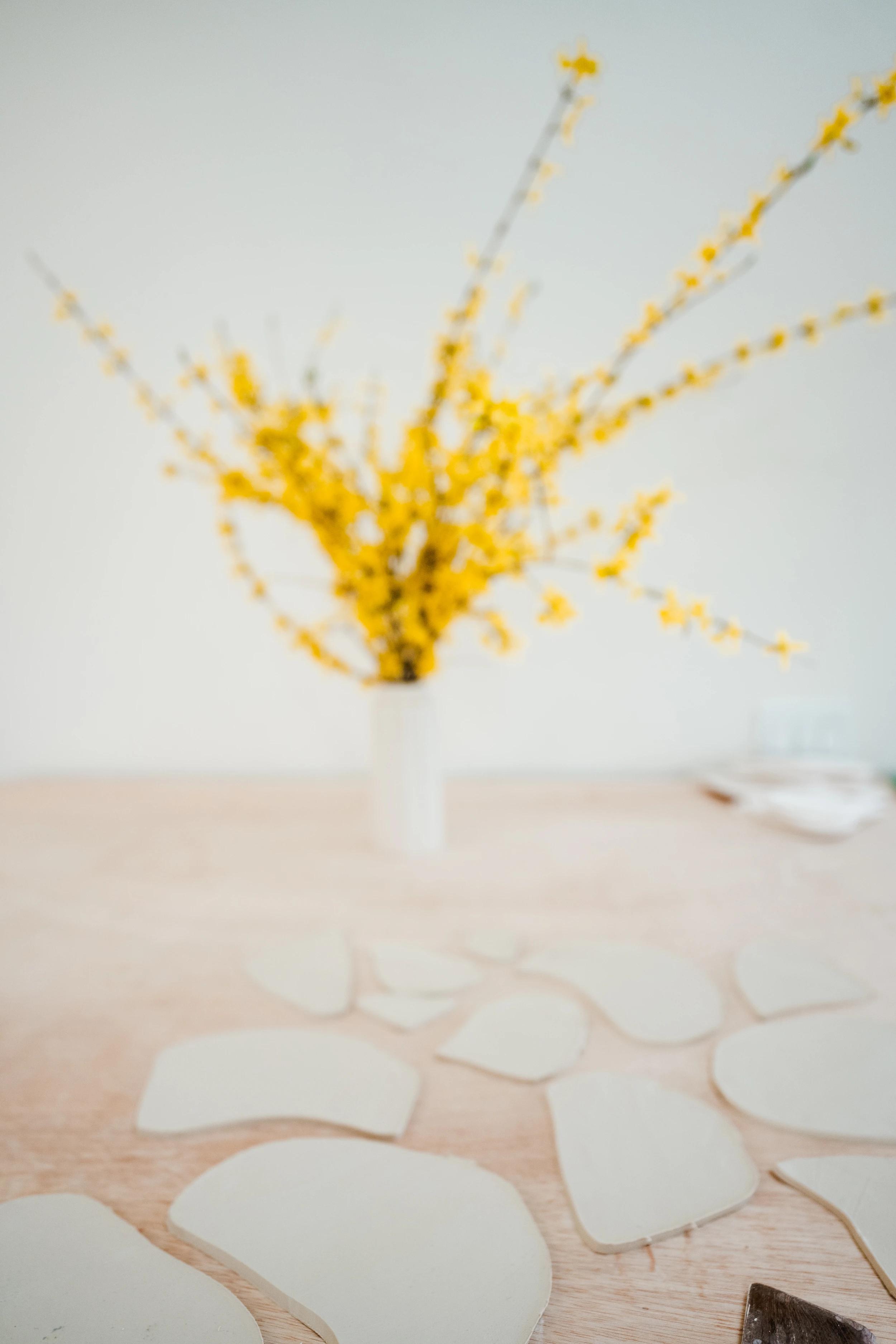 Irregularly-shaped clay pieces on a wooden table next to a vase of yellow flowers in artist Christina Watka's studio. 