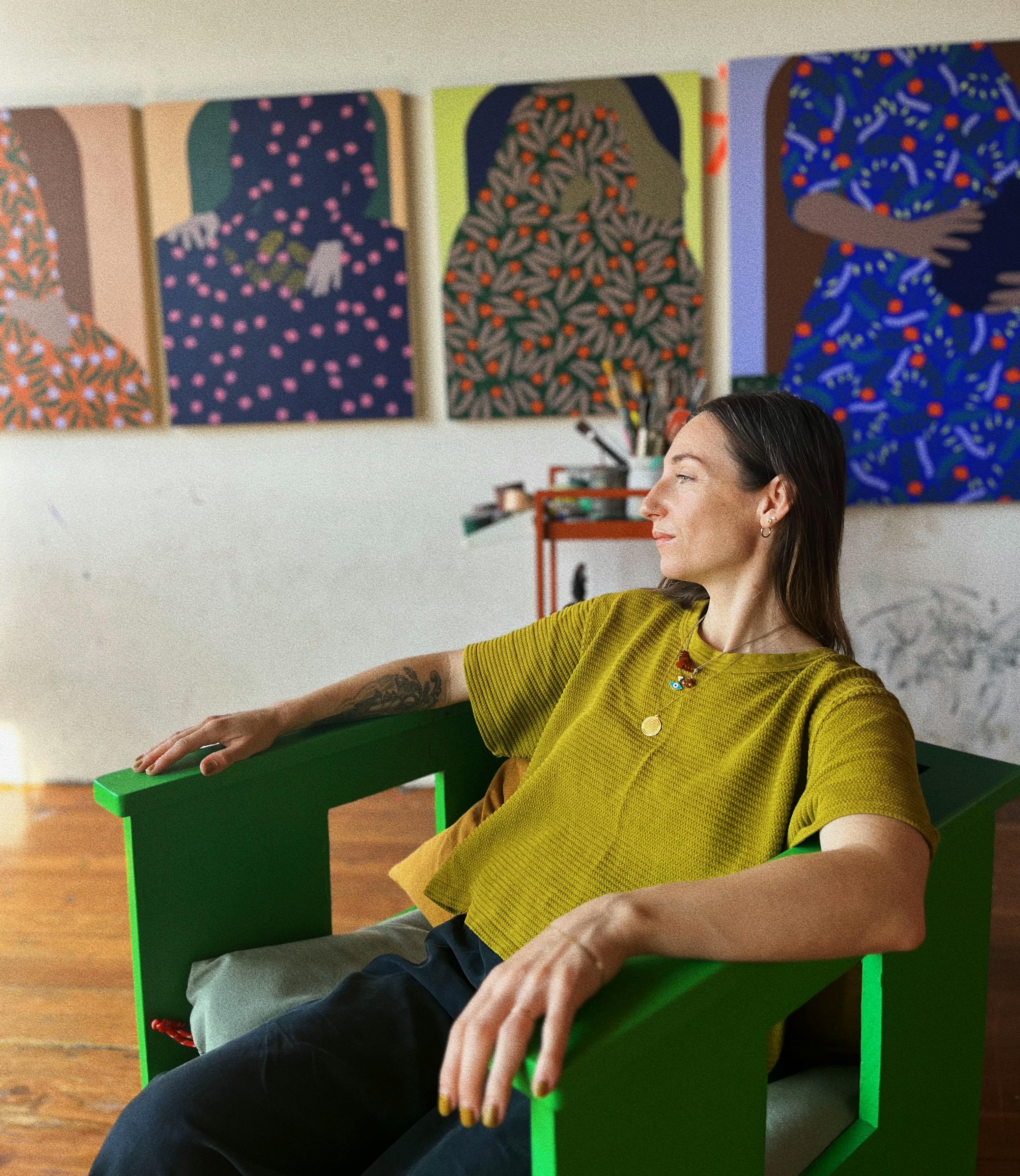 Artist Carmen McNall sitting in a green chair in her studio with her figurative paintings installed on a white wall behind her.