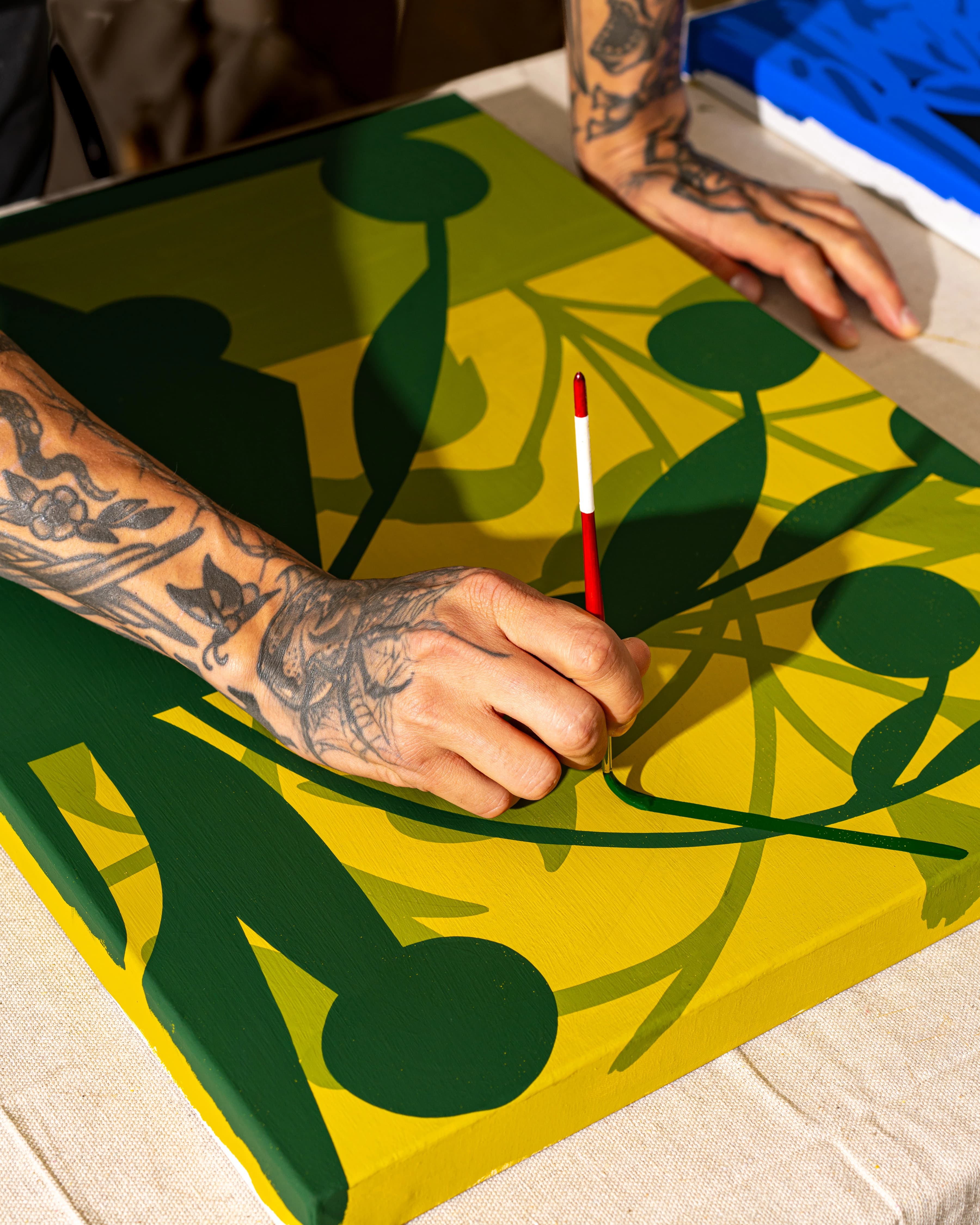 Close-up of artist Eddie Perrote using a thin paintbrush to paint green leaves on yellow canvas during his Uprise Art x MacArthur Place Residency.