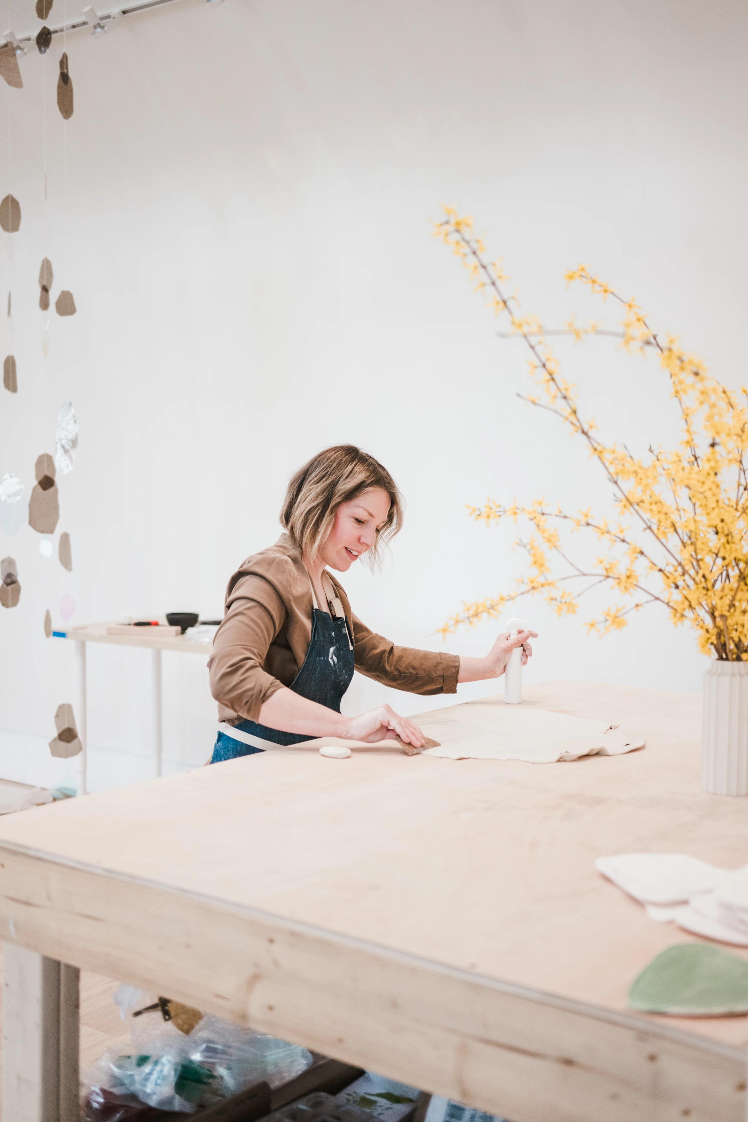 Artist Christina Watka working on clay discs at a large wooden table with yellow flowers in her studio.