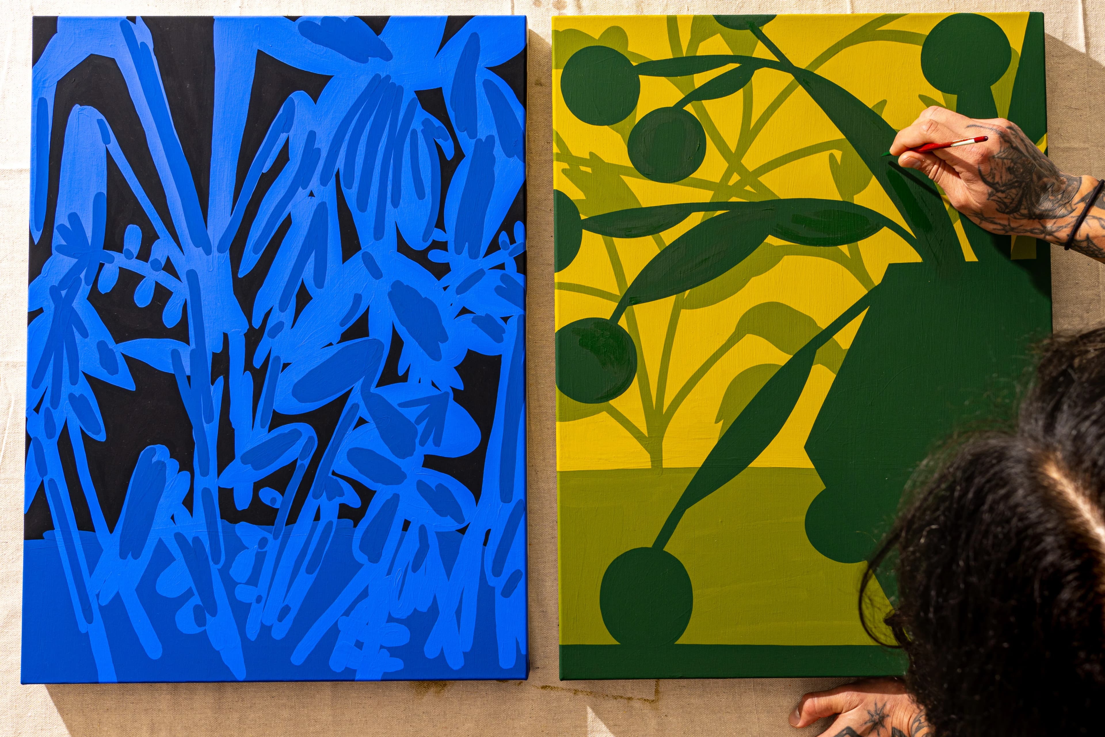 Aerial view of artist Eddie Perrote painting two vivid, botanical paintings, one blue and one yellow and green, during his Uprise Art x MacArthur Place Residency.