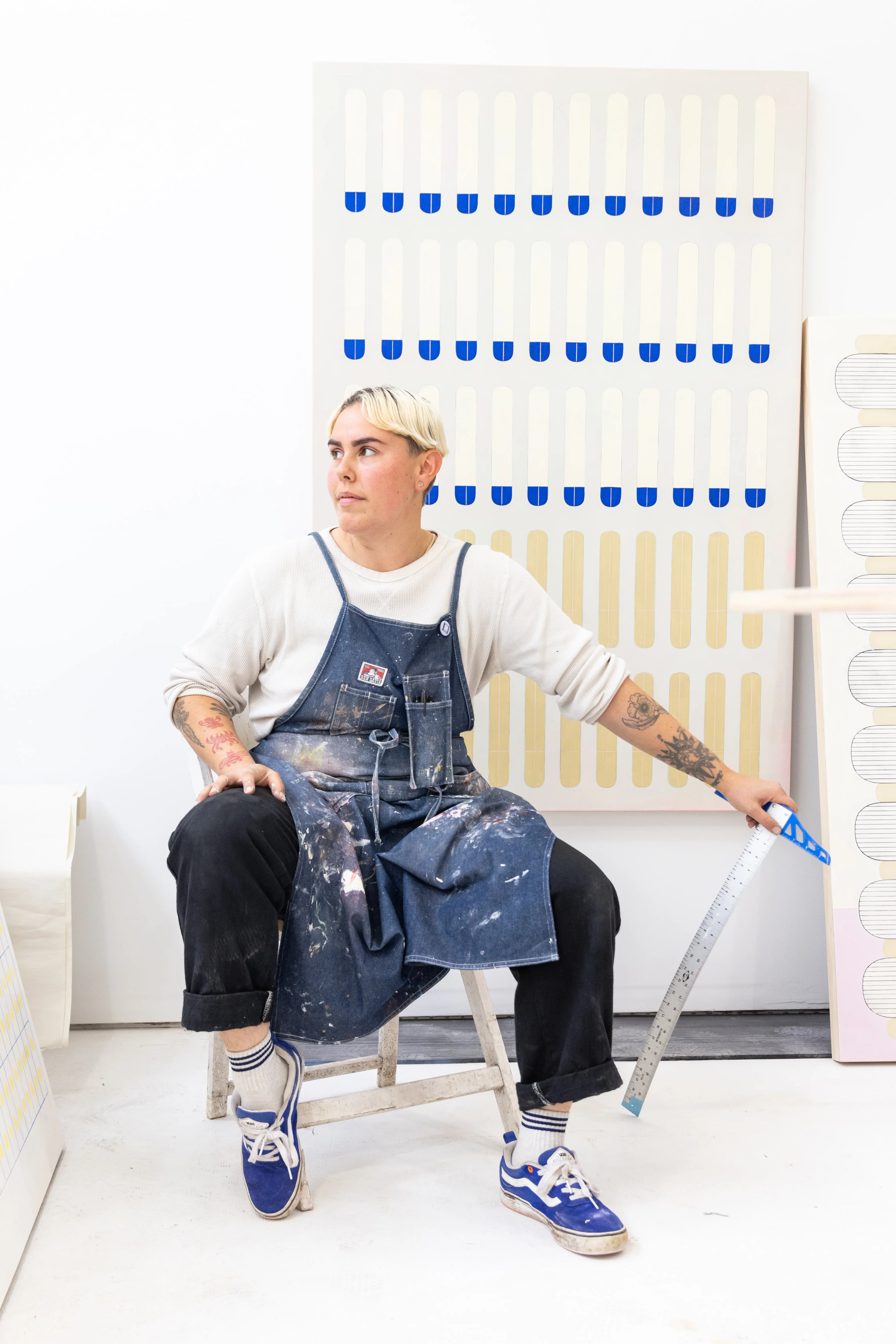 Artist Arielle Zamora sitting in their studio in front of a large, blue and yellow patterned work installed on a white wall.