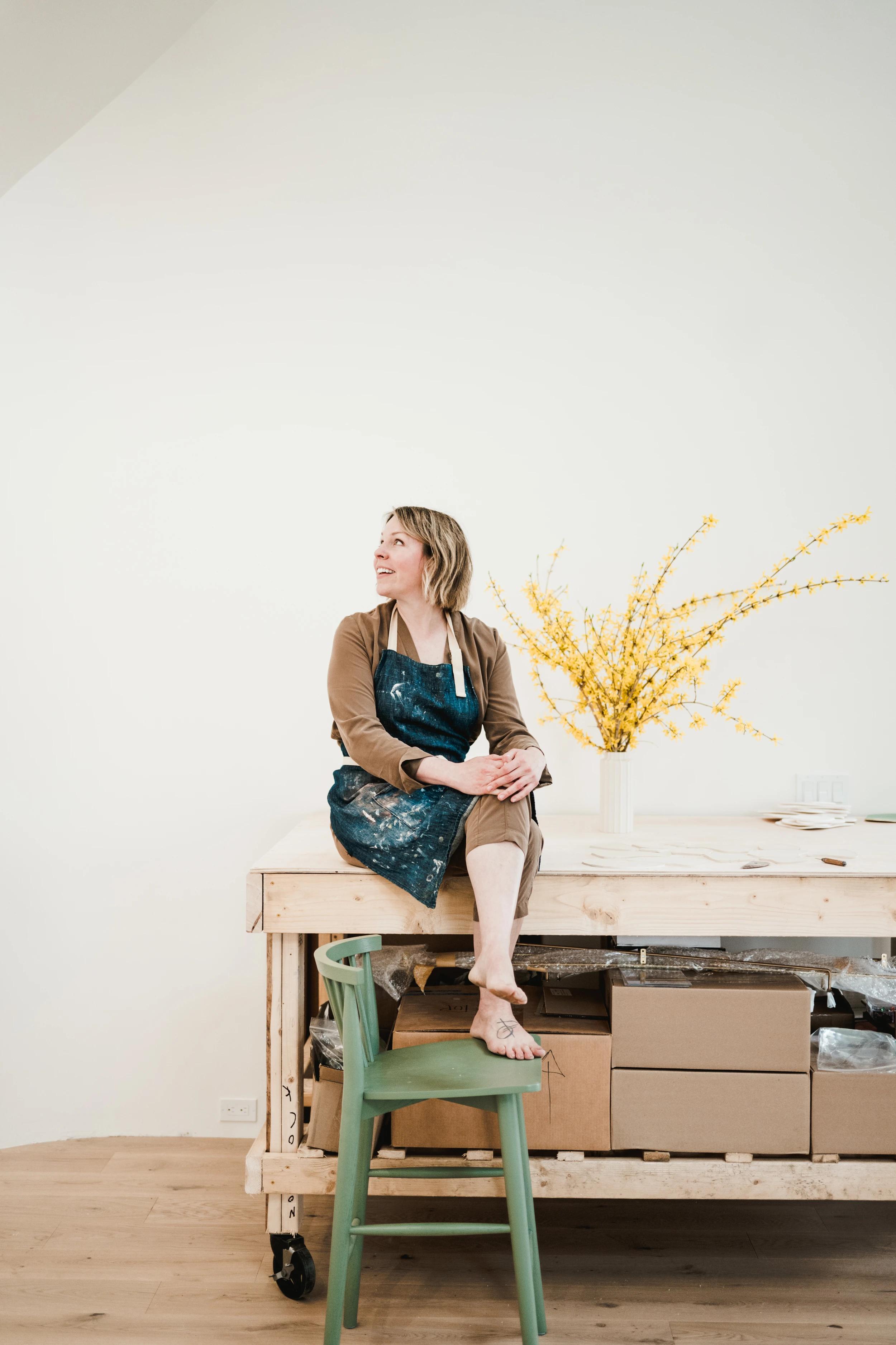 Artist Christina Watka sitting on a large wooden workbench with her feet on a green chair in her Maine studio.