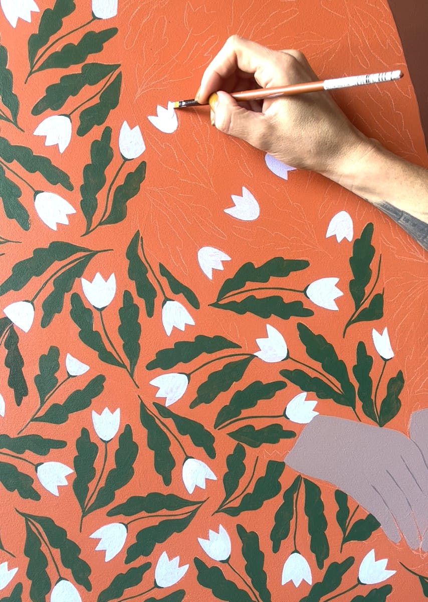 Close-up of artist Carmen McNall painting a white floral pattern on an orange wood panel.