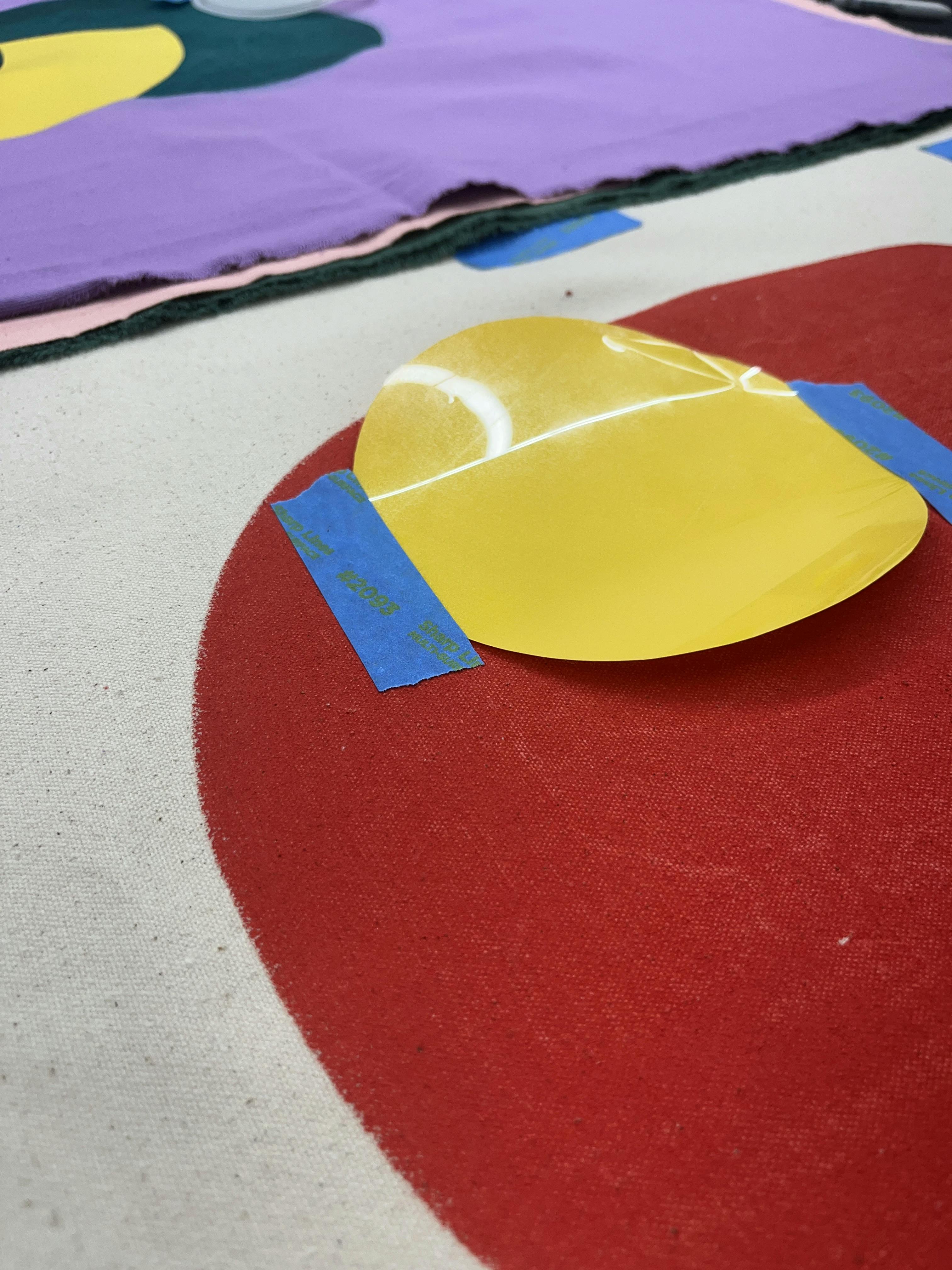 An in-progress work by artist Chad Kouri of a yellow circle taped to red fabric.
