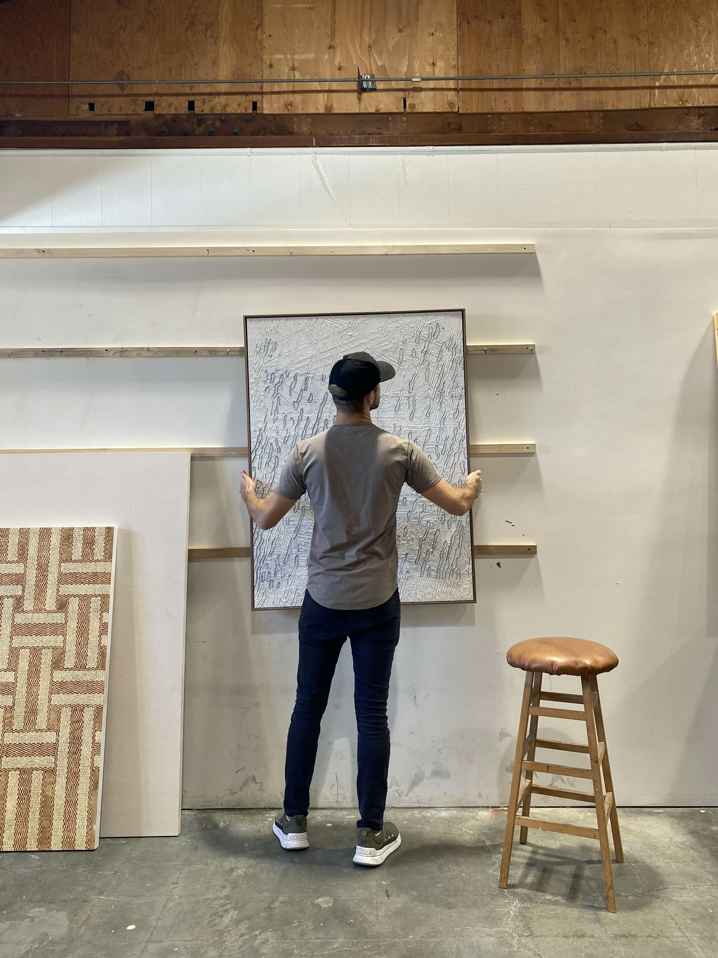 Artist Blake Aaseby in his studio carrying a large, gray textured painting.